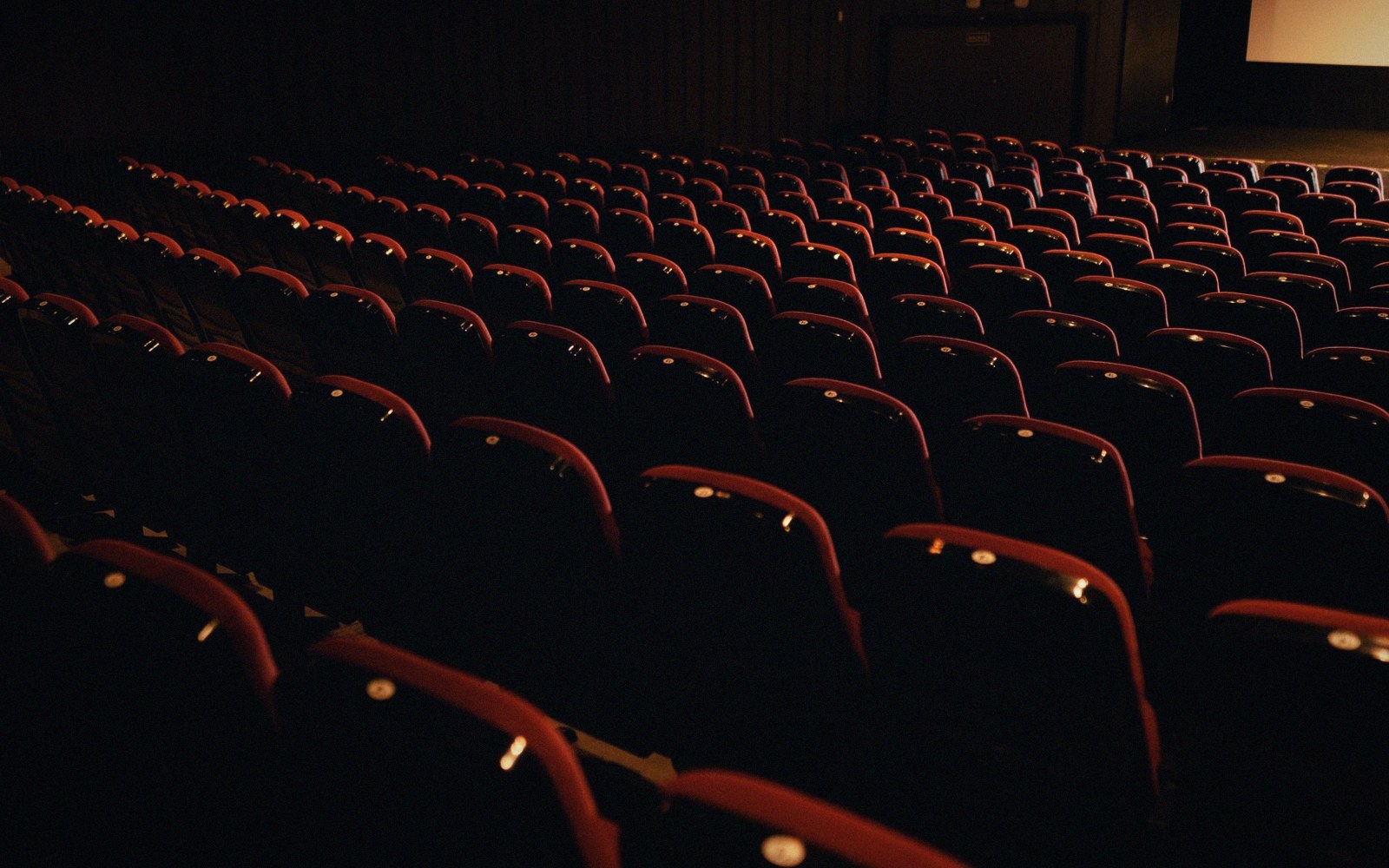 Rows of empty red cinema seats viewed from the aisle, house lights up, screen dark, before a screening begins