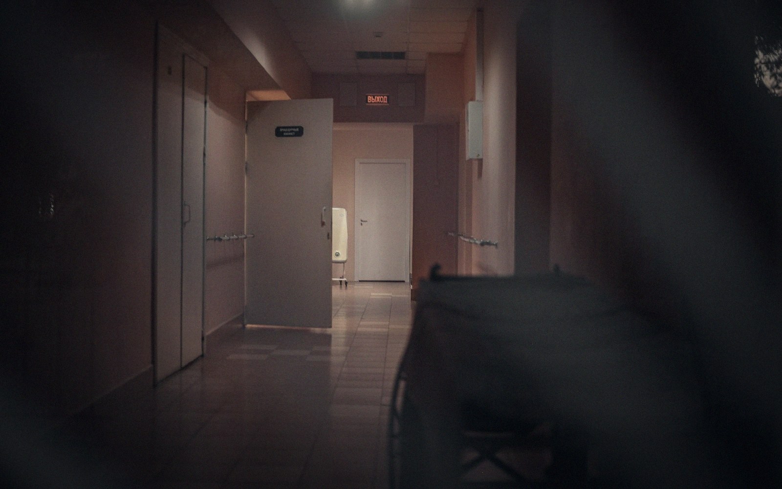 A dimly lit hospital corridor with white doors and an illuminated exit sign