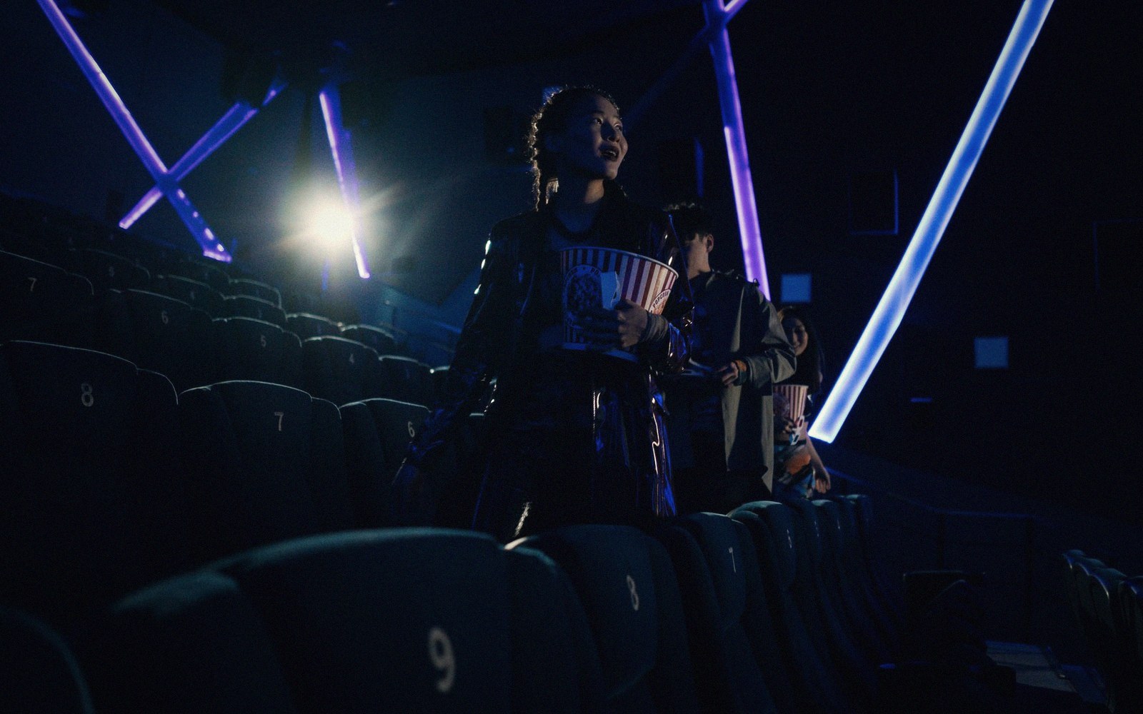 Moviegoers carry popcorn buckets down the aisle of a dark theater lit by neon beams