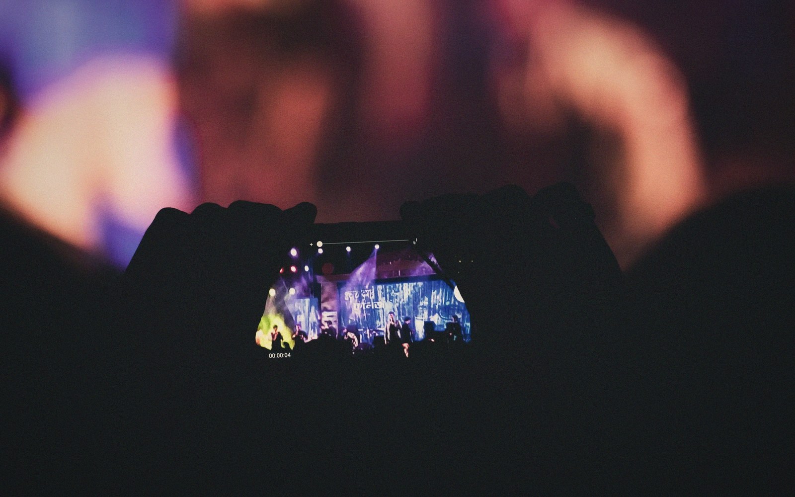 Silhouetted hands hold up a phone recording a concert stage from the crowd