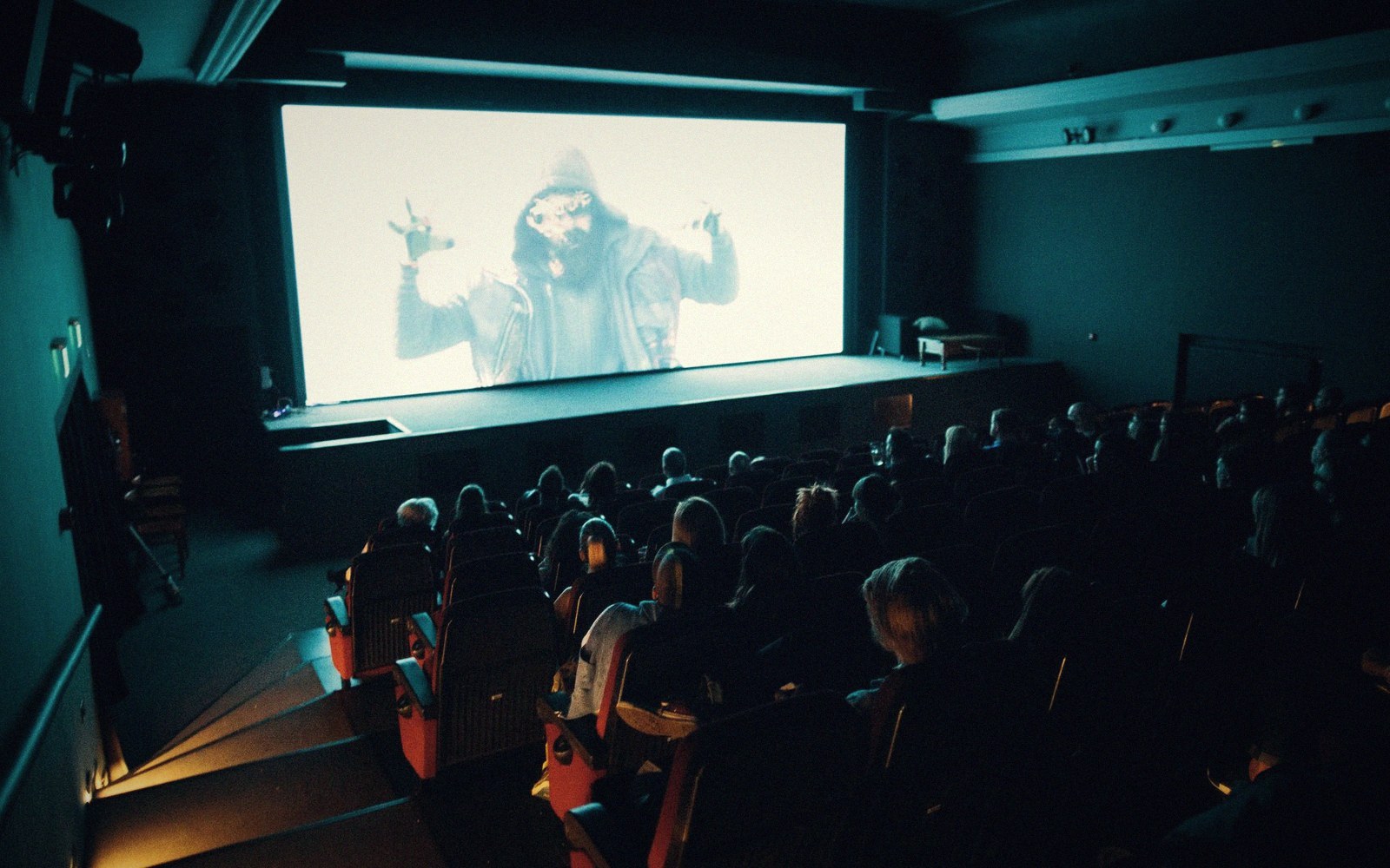 Audience members watch a hooded figure on screen in a darkened cinema auditorium