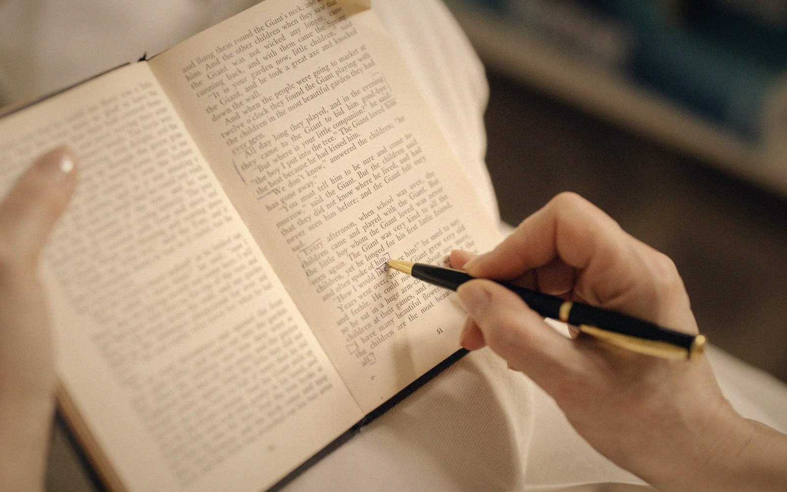 Hands hold an open book while marking a line of printed text with a pen