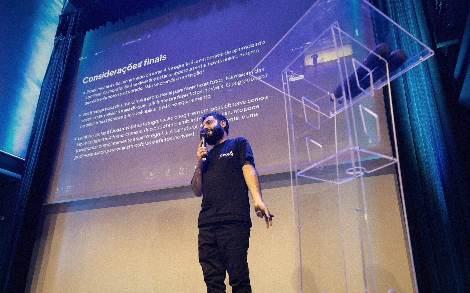 A speaker gestures while presenting slides with Portuguese text on stage