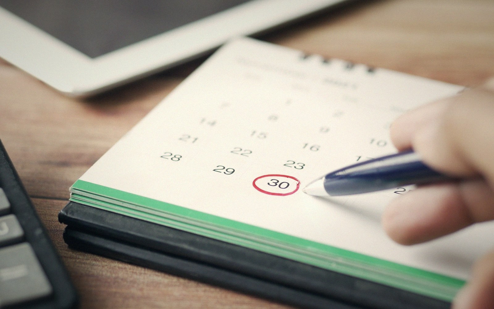 A hand circles the date 30 in red pen on a desk calendar