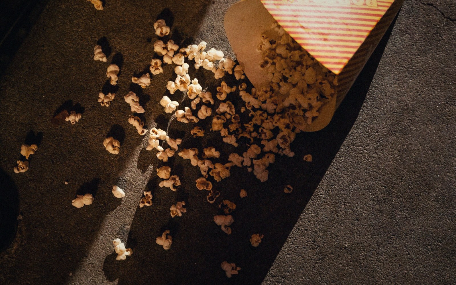 A striped popcorn box tipped over, spilling kernels onto pavement in dim light